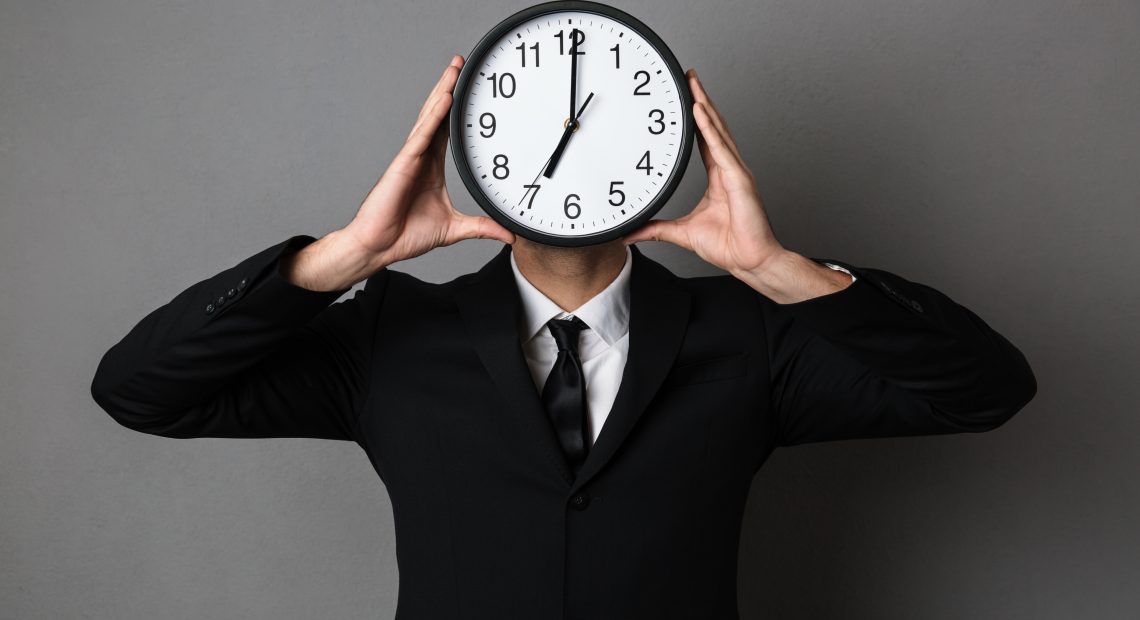 Young man in black suit holding clock in front of his face, isolated on gray background